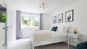 A white bedroom with a bed, a bedside table, a window with curtains, and framed pictures on the wall. - Coastal Retreat for 10 with Hot Tub (West Wittering)