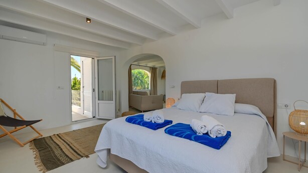 A bedroom with a large bed, a chair, a small table, and a view of the outdoors.