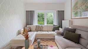 Uma sala de estar com um sofá, uma mesa de centro e um vaso com flores secas. Há uma janela com cortinas, um radiador e um mapa emoldurado na parede.