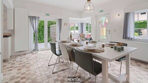 Uma sala de jantar com uma mesa branca posta para quatro, cadeiras, um lustre e vista para o exterior através de grandes janelas.