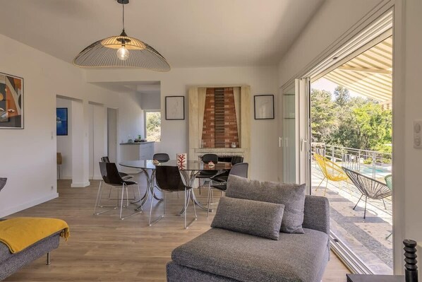 A modern living room with a grey sofa, dining area with chairs, and a large window overlooking a pool. - Villa Jeanne (Linguizzetta)
