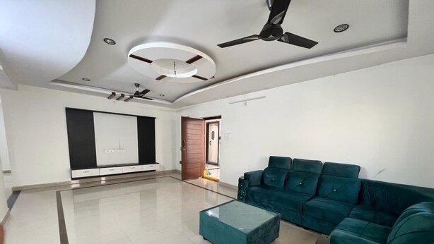 A spacious living room with a teal sofa set, a glass coffee table, and a ceiling fan. - The Excellency Apartments (Hyderabad)