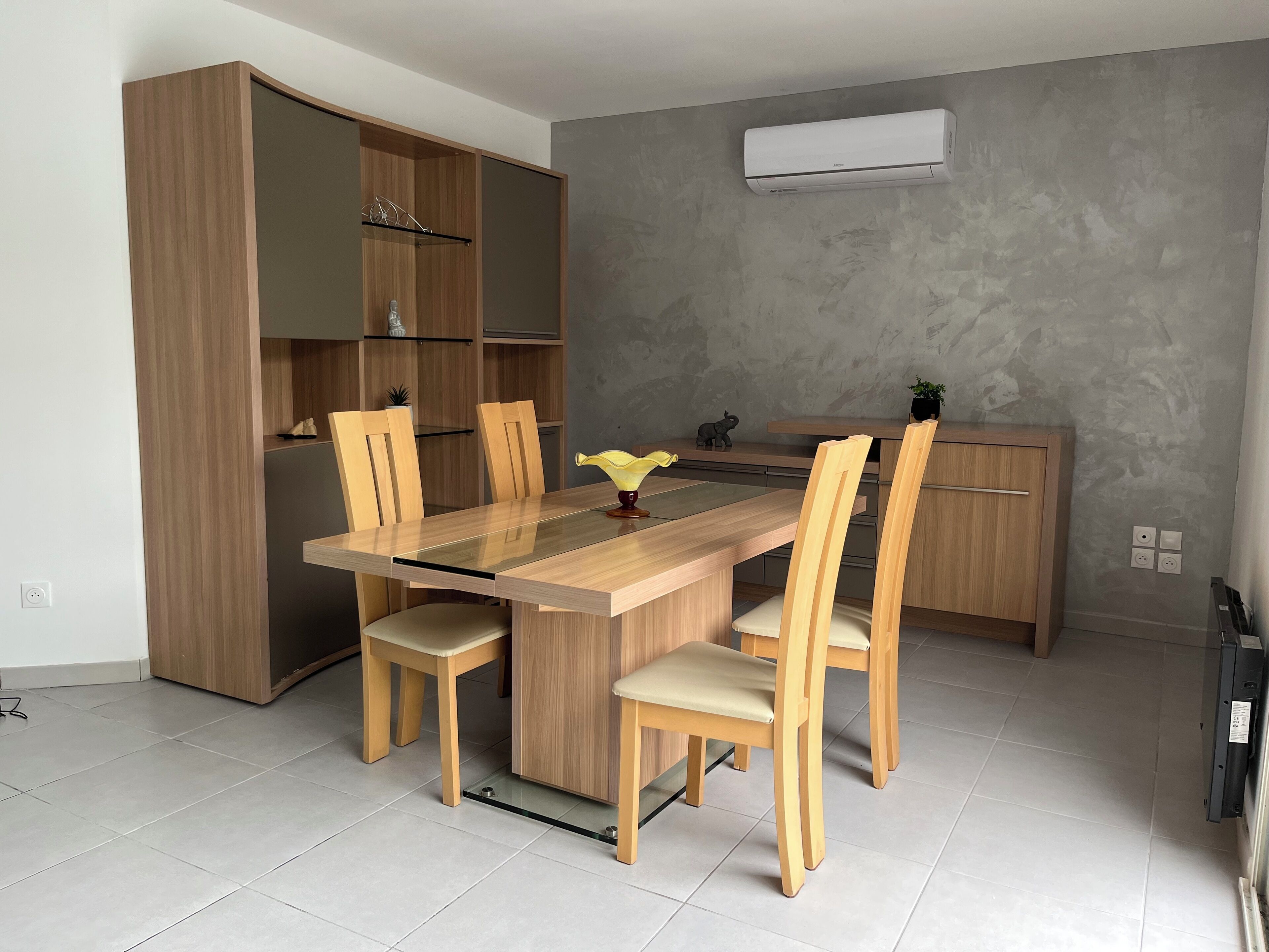 A modern kitchen with a wooden dining table, four chairs, a built-in cabinet, and an air conditioning unit on the wall.