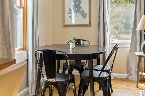 Une table de salle à manger ronde, entourée de quatre chaises noires, avec une plante en pot et une œuvre d’art encadrée accrochée au mur.