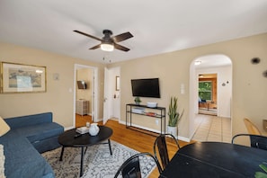 A living room with a blue sofa, a coffee table, a TV on a black stand, and a ceiling fan. - Cozy 2BR Loft, Stroll to Downtown A2 (Ann Arbor)