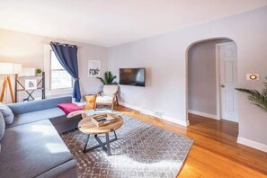 A modern living room with a sofa, a coffee table, a TV, and a door leading to another room. - Large 4BR Home, Walk to Downtown A2 (Ann Arbor)