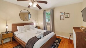 A hotel room with a bed, a ceiling fan, a bedside table with a lamp, a mirror, a window with curtains, and a desk with a chair. - Large 4BR Home, Walk to Downtown A2 (Ann Arbor)
