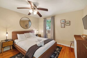 A hotel room with a bed, a ceiling fan, a round mirror, a bedside table with a lamp, a window with curtains, and a desk with a chair. - Large 4BR Home, Walk to Downtown A2 (Ann Arbor)