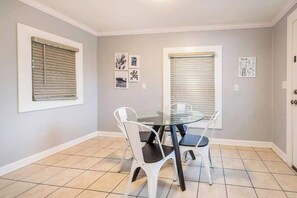 A dining area with a glass table and white chairs, two windows with blinds, and wall art. - Large 4BR Home, Walk to Downtown A2 (Ann Arbor)