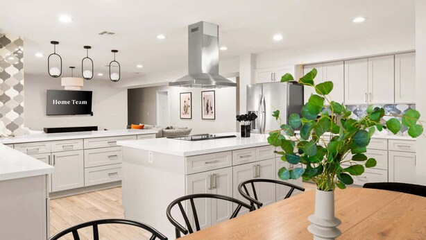 Uma cozinha moderna com armários brancos, uma ilha central, iluminação pendente e uma área de jantar com mesa e cadeiras de madeira.