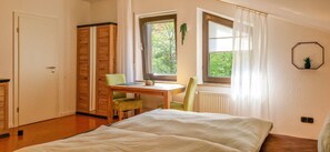 A bedroom with a bed, a desk, and a chair. There is a window with curtains, a radiator, and a door.