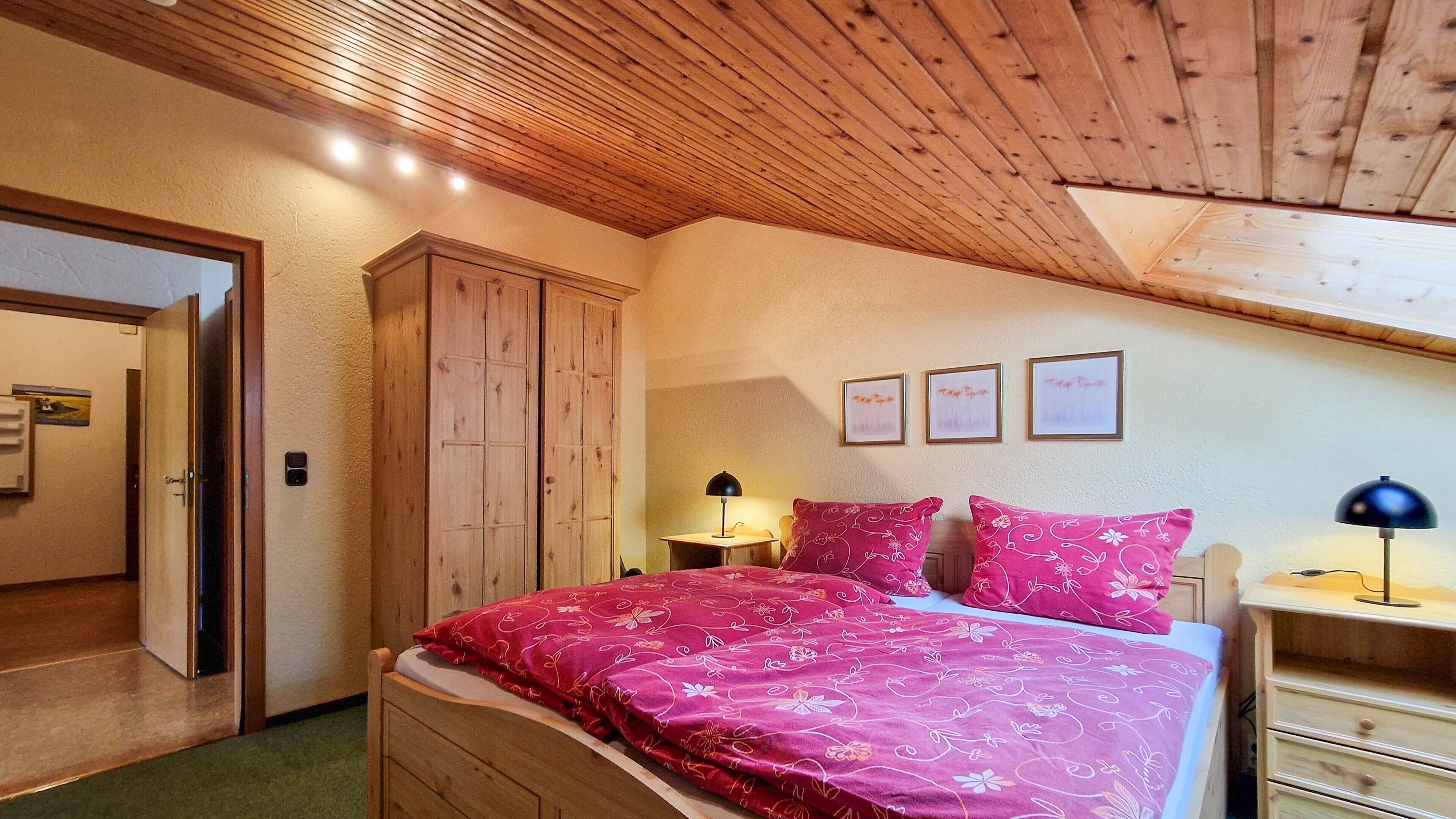 Ein Schlafzimmer mit Holzboden, einem Bett mit floralem Bettzeug, einem Nachttisch, einer Kommode und einer Tür zu einem anderen Raum.