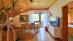 A room with a wooden ceiling, a dining table with chairs, a TV on a stand, and a window with curtains.