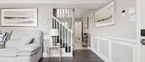 A living room with a grey sofa, a small table with a lamp, and a staircase leading to another room.