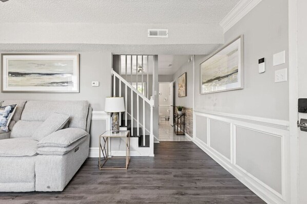 A living room with a grey sofa, a small table with a lamp, and a staircase leading to another room.