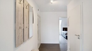 A hallway with a wooden wall panel, a door, and a glimpse of a bedroom with a bed and a window.