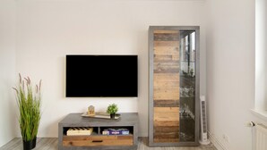 A modern living room with a flat-screen TV mounted on the wall, a wooden sideboard with a glass cabinet, and a potted plant.