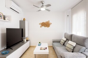 Ein modernes Wohnzimmer mit Flachbildfernseher, Deckenventilator, grauem Sofa, weißem Couchtisch und einem Wandbild.