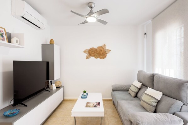 A modern living room with a flat-screen TV, a ceiling fan, a grey sofa, a white coffee table, and a decorative wall piece.
