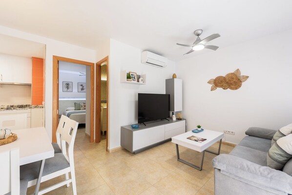 Un salon moderne avec un téléviseur, des meubles blancs, un ventilateur de plafond et une cuisine visible au fond.