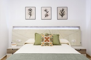 Une chambre à coucher comprenant un lit, deux tables de chevet, une tête de lit et trois reproductions botaniques encadrées accrochées au mur.