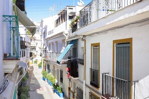 Une rue étroite bordée d’immeubles à plusieurs étages, de balcons ornés de plantes en pot et surmontée d’un auvent bleu.