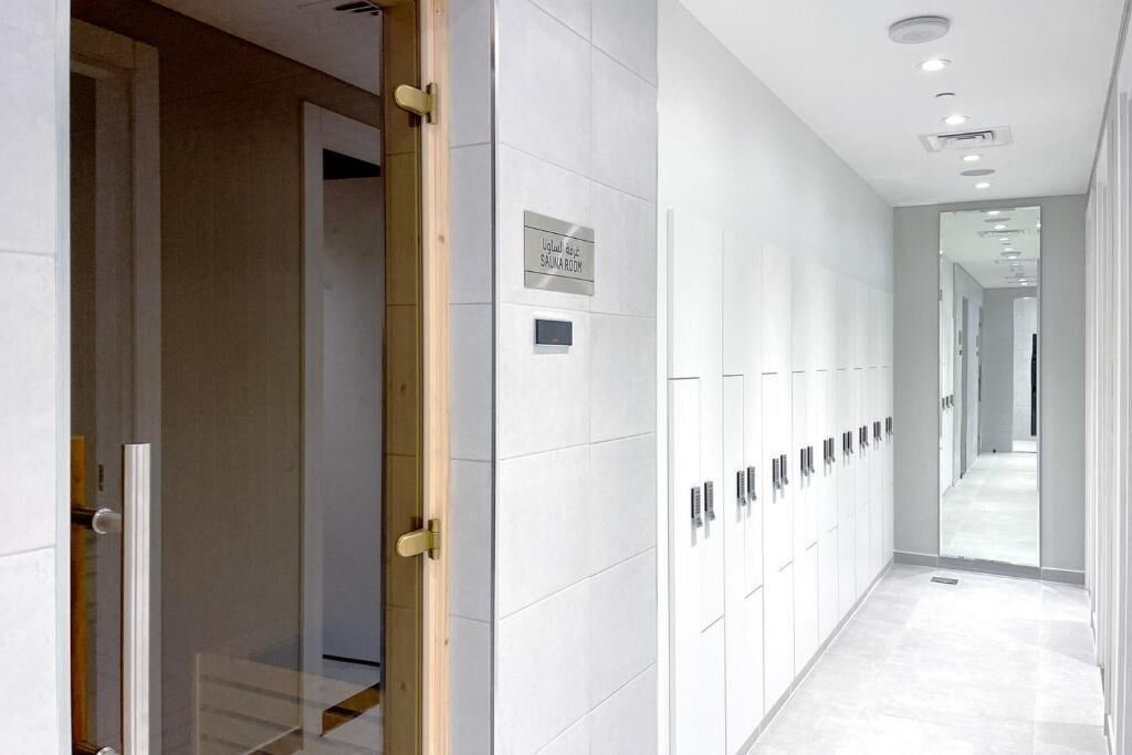 A modern hallway with lockers, a sign indicating a sauna room, and a mirrored wall reflecting the corridor.