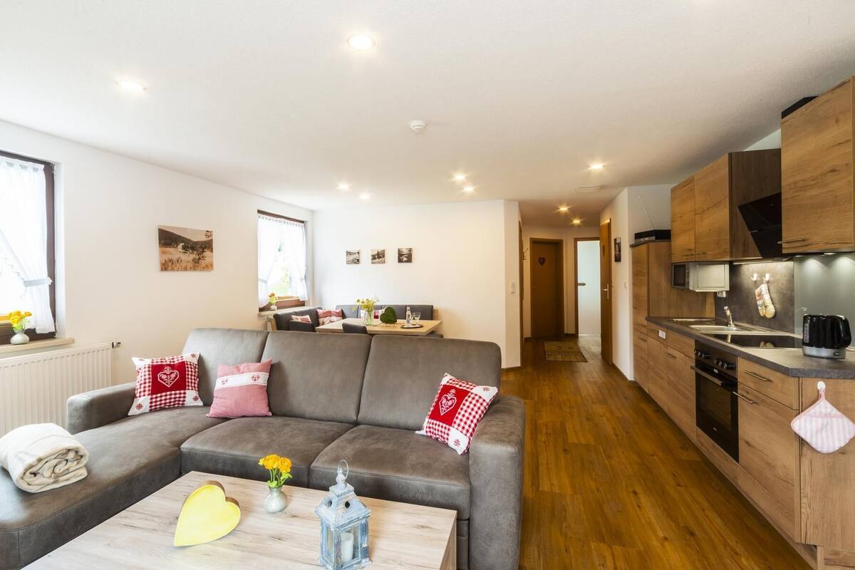 A modern living room with a grey sofa, a wooden coffee table, and a kitchen with wooden cabinets and a microwave.