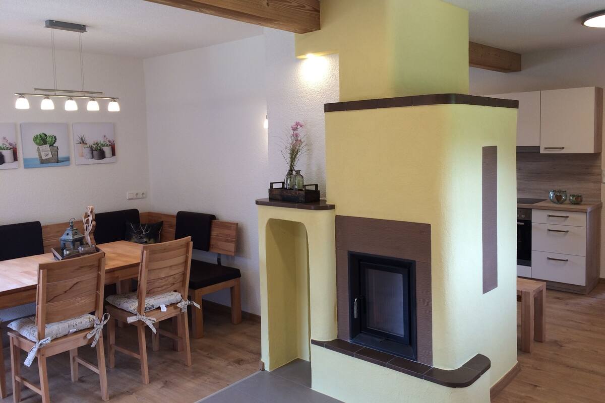 A dining area with a wooden table and chairs, a built-in fireplace, and a kitchen in the background.