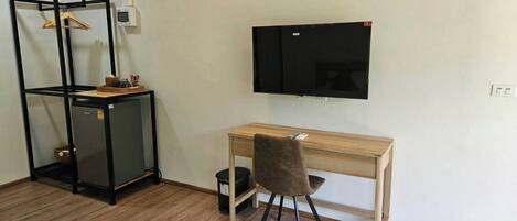 A room with a wooden desk, a chair, a TV mounted on the wall, and a black shelving unit with a basket on top.