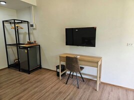 Una habitación con un escritorio de madera, una silla, un televisor montado en la pared y un estante negro con una cesta encima.