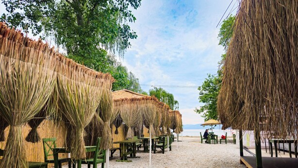 Un restaurante frente a la playa con sombrillas de paja y mesas de madera.