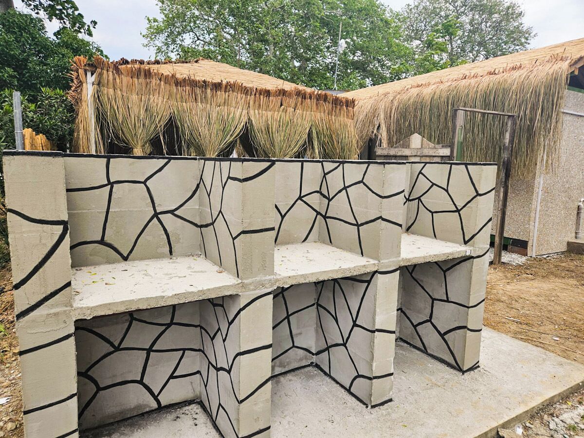 Un banc en béton orné d’un motif géométrique, placé devant une structure à toit de chaume et entouré de verdure.