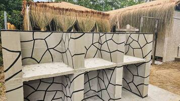 Un banc en béton orné d’un motif géométrique, placé devant une structure à toit de chaume et entouré de verdure.