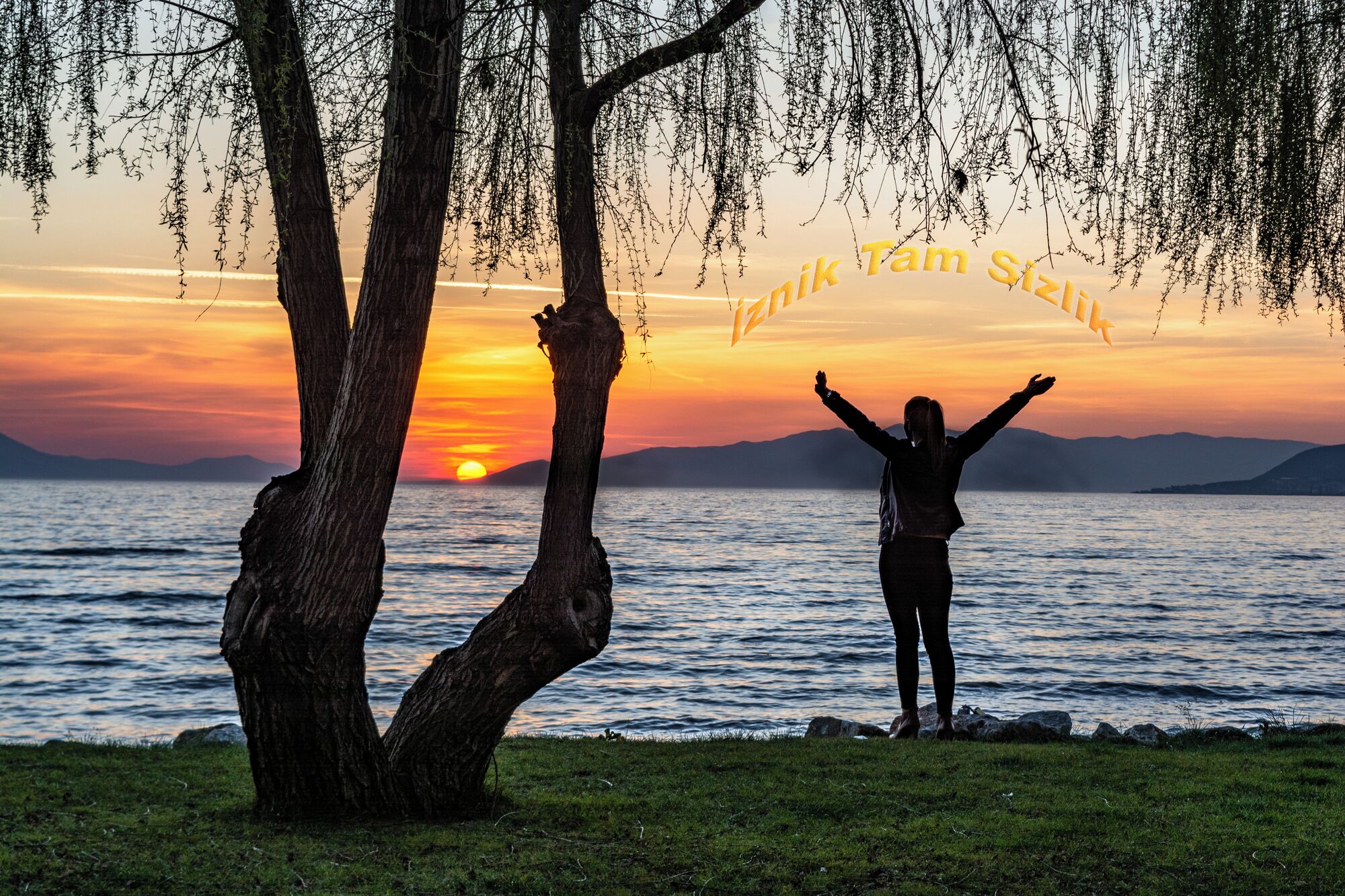 A person standing by a lake at sunset with trees and mountains in the background.