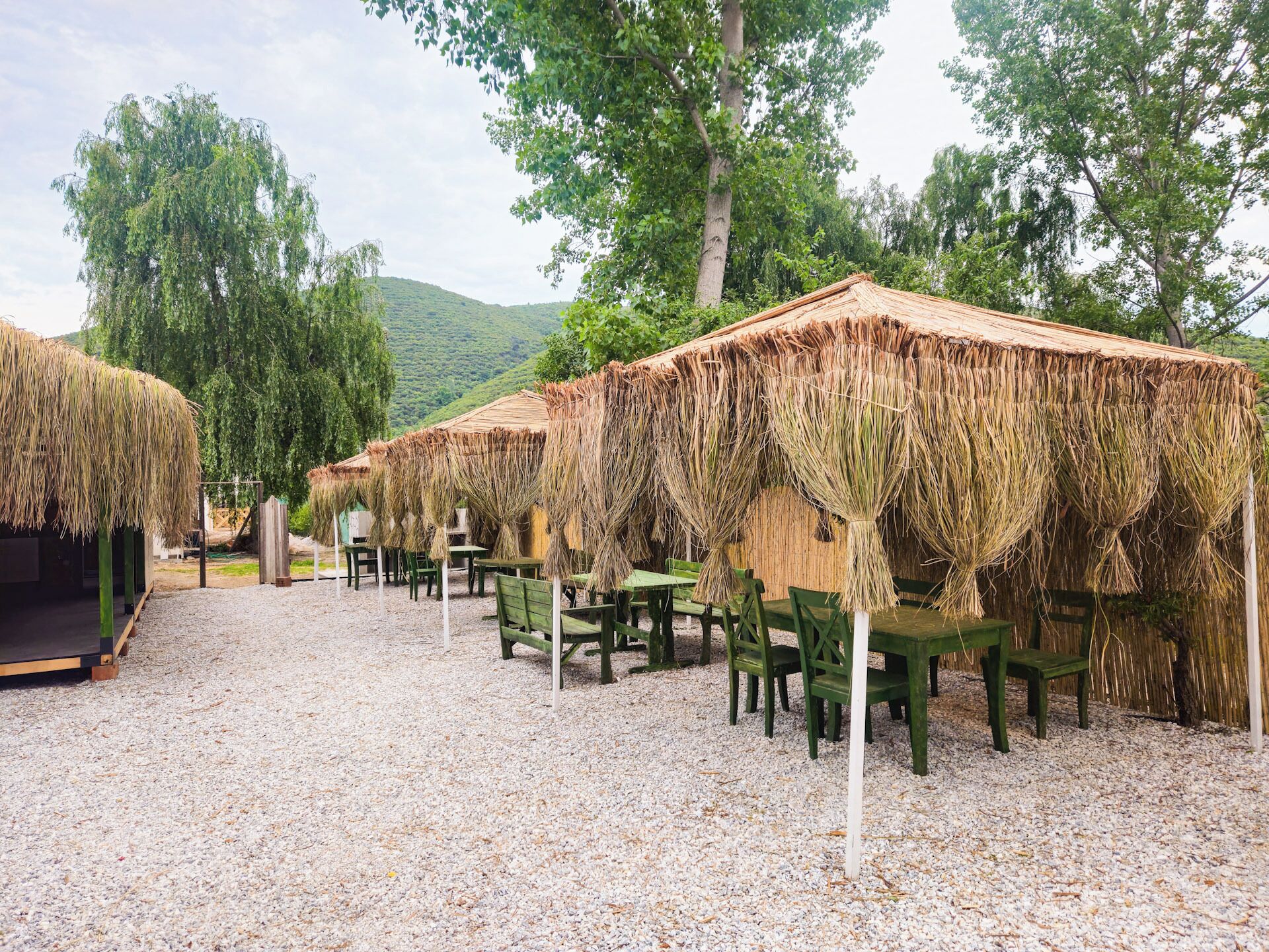 Filas de cabañas con techo de paja, sillas y mesas verdes, sobre un terreno de grava, con árboles y colinas al fondo.
