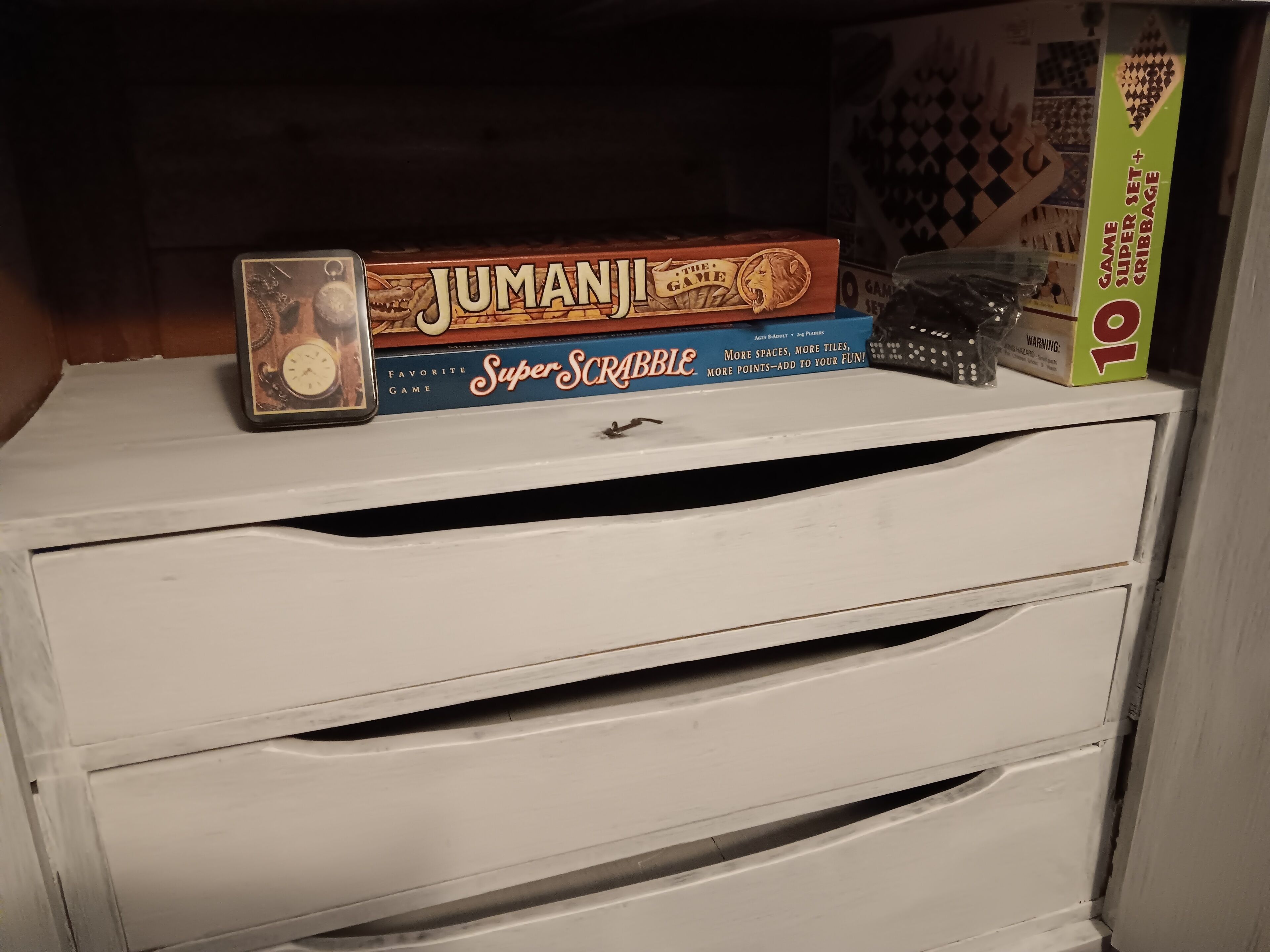 Eine Spielbox mit den Aufschriften "Jumanji" und "Super Scrabble" auf einer weißen Schublade.