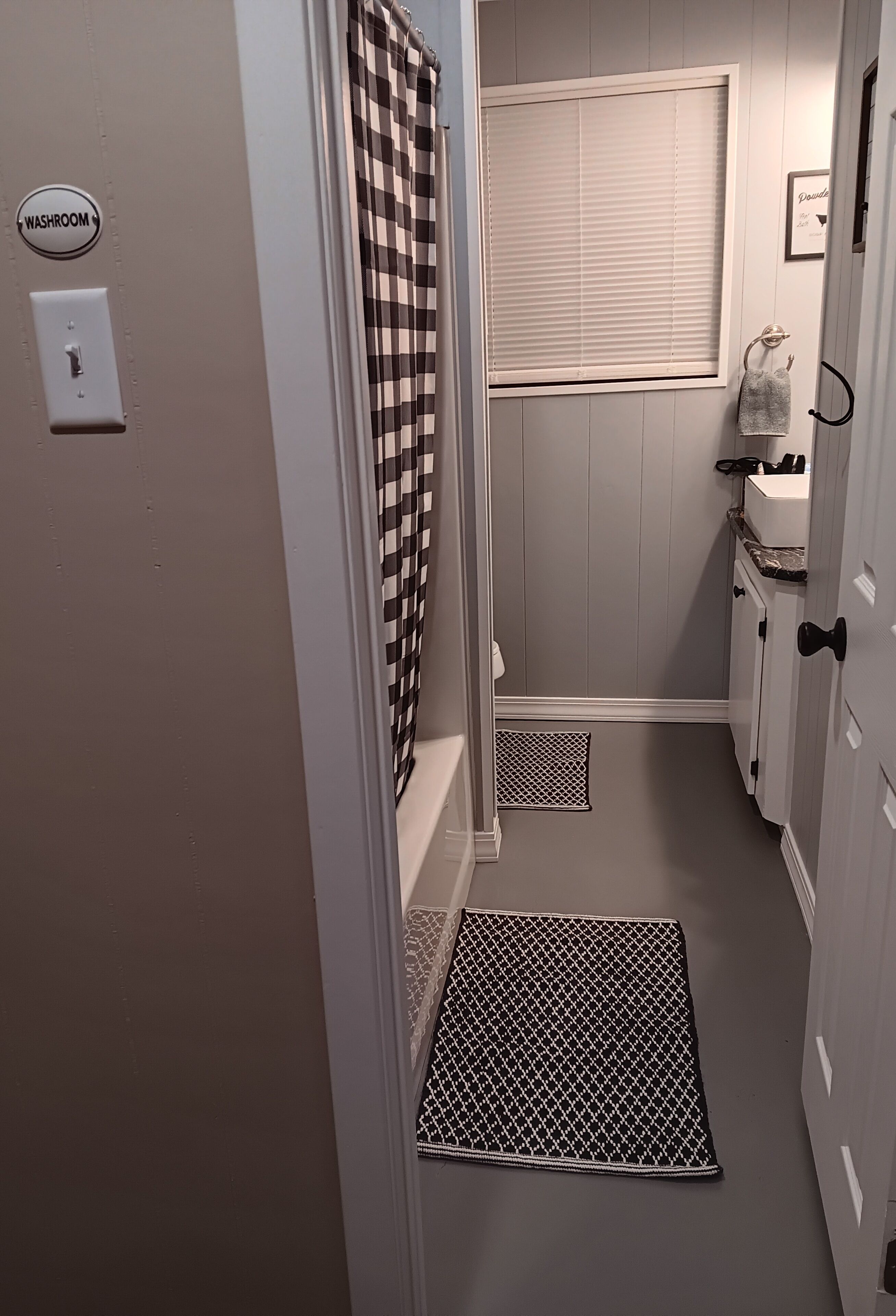 A small, narrow bathroom with a checkered shower curtain, a window with blinds, a towel rack, and a sign indicating a washroom.