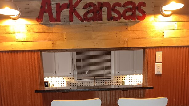 Un coin bar avec deux tabourets blancs, un mur en bois orné d’une enseigne indiquant « Arkansas », et une fenêtre avec des stores.