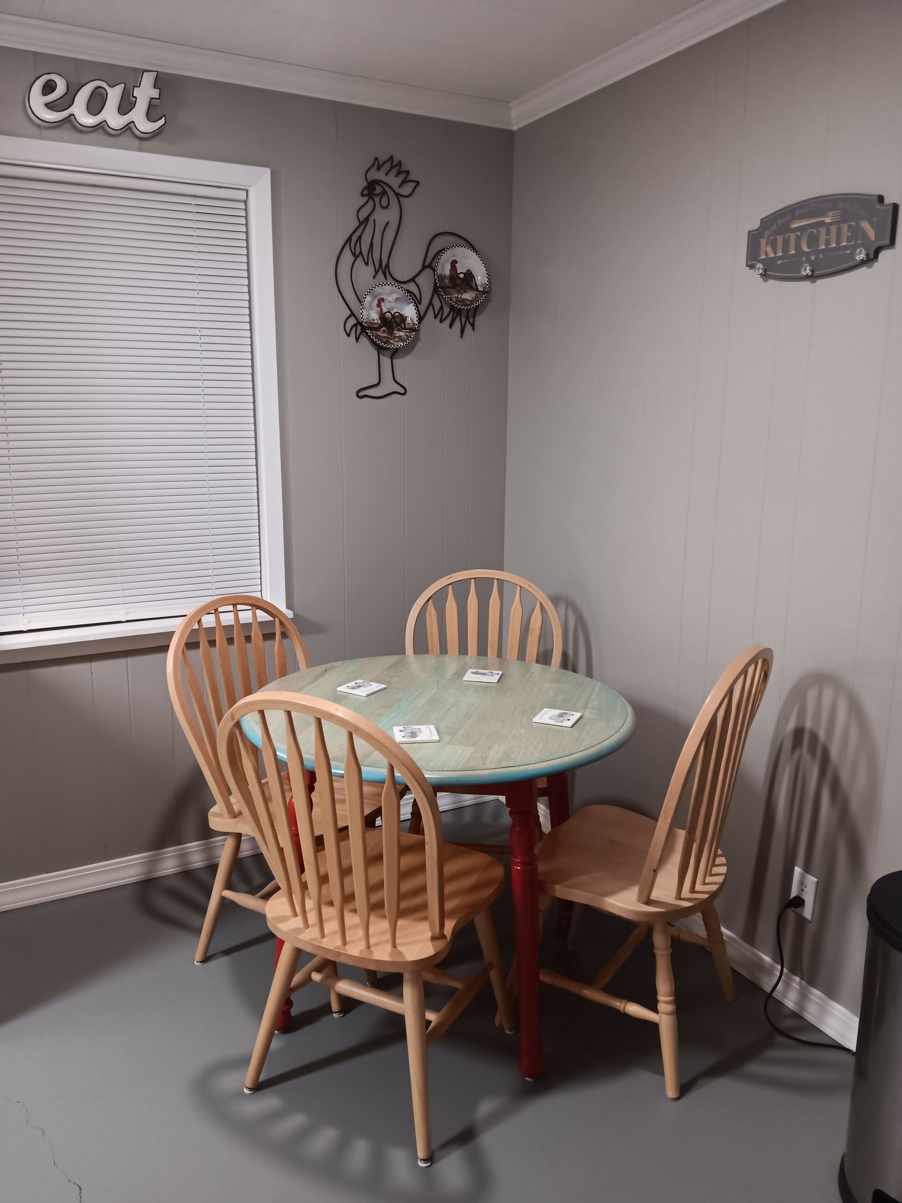 A dining area with a round table and four wooden chairs, a rooster wall decoration, and a 'KITCHEN' sign on the wall.