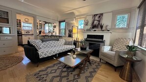 A spacious living room with a sofa, coffee table, and armchair. A fireplace and a dining area are visible in the background. - Beautiful Normal Heights Living (San Diego)