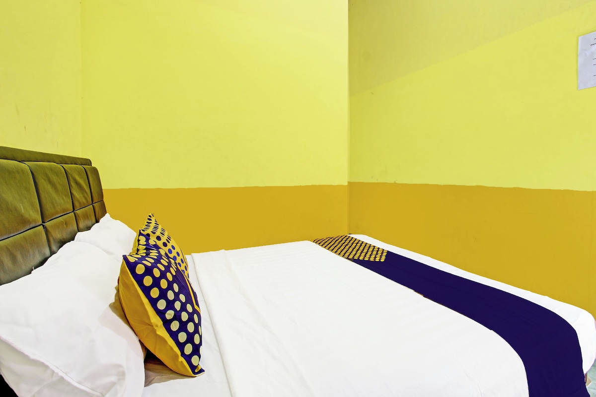 Una cama con sábanas blancas y almohada azul y amarilla, frente a una pared amarilla y un cabecero marrón.