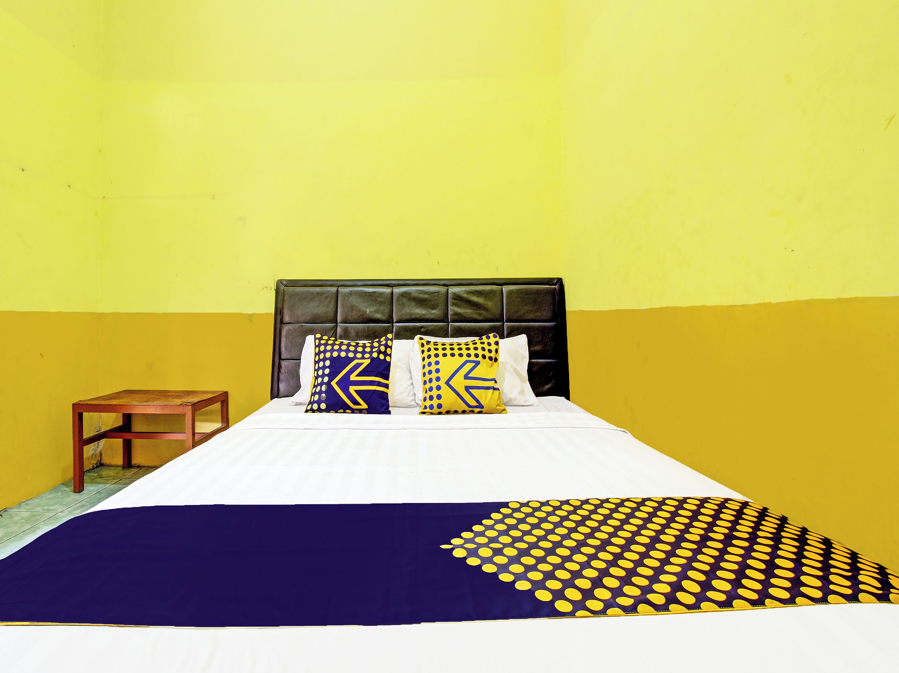 Una cama con cabecera negra, dos almohadas con diseños azules y amarillos, una mesita de noche de madera y una pared blanca y amarilla.
