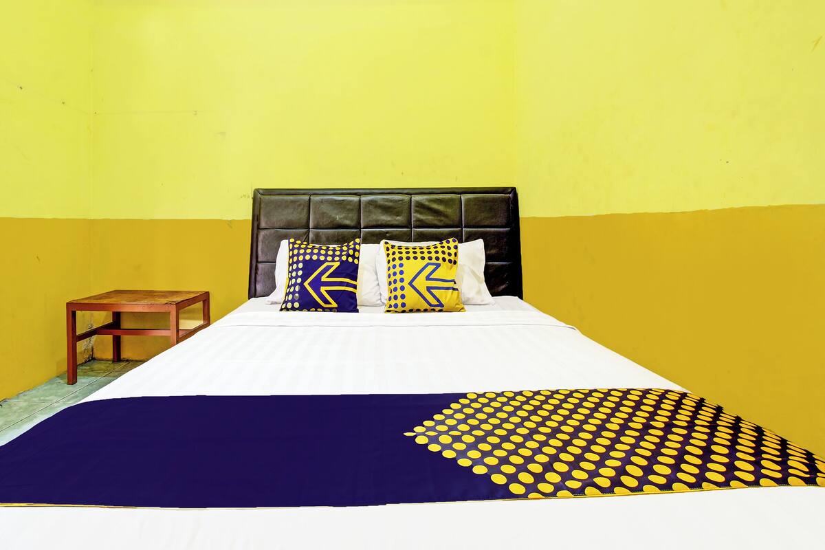 Una cama con cabecera negra, dos almohadas con diseños azules y amarillos, una mesita de noche de madera y una pared blanca y amarilla.