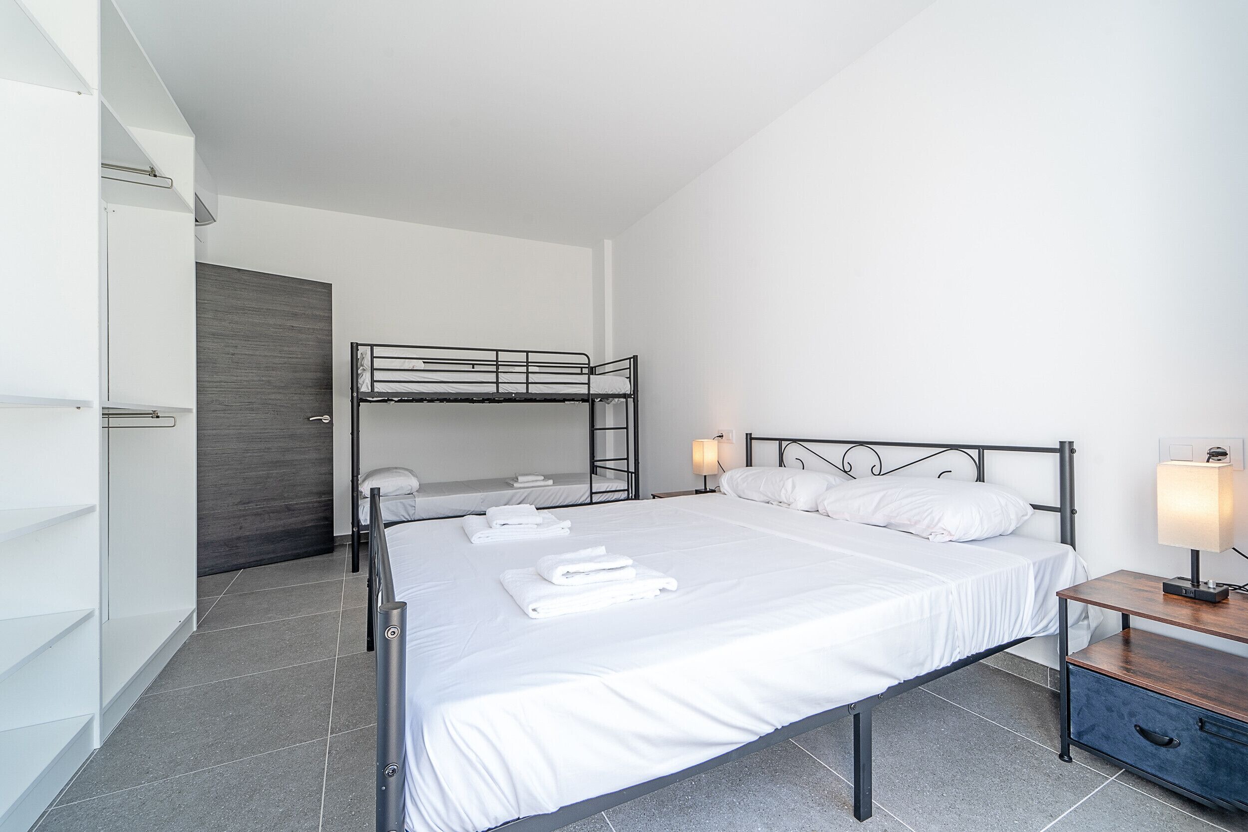 Una habitación de hotel moderna con una cama grande, una cama nido más pequeña, una mesita de noche con lámpara y un armario.