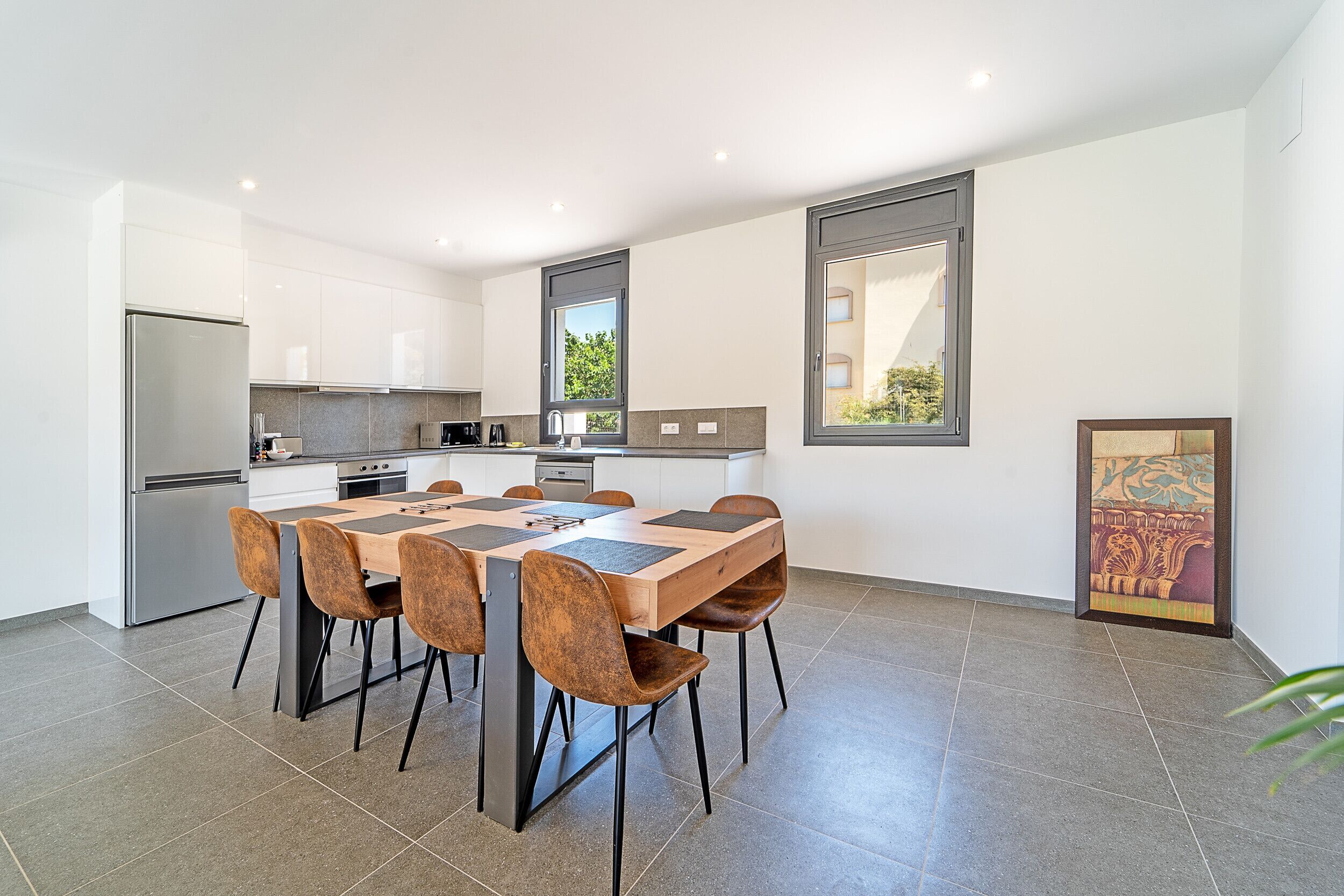 A modern kitchen with a dining area, featuring a large table and chairs, a refrigerator, and a window with a view of greenery.