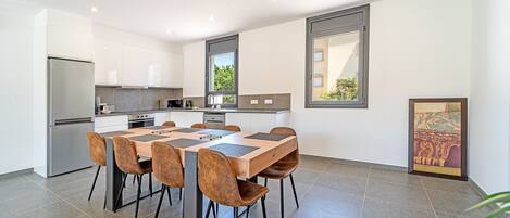 A modern kitchen with a dining area, featuring a large table and chairs, a refrigerator, and a window with a view of greenery.