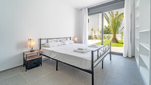 A modern bedroom with a metal bed frame, white linens, a bedside table with a lamp, and a view of a green landscape through large windows. - WELCS VILLA 167 EMP con piscina privada (Empuriabrava)