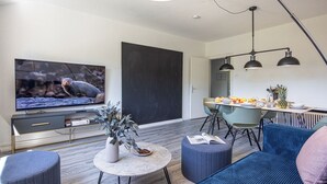 En moderne stue med fjernsyn, sofa, skammel og spiseområde med stole og bord.