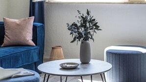 Et værelse med en blå sofa, et rundt bord med en vase og en skål, samt et vindue med udsigt over grønne områder.